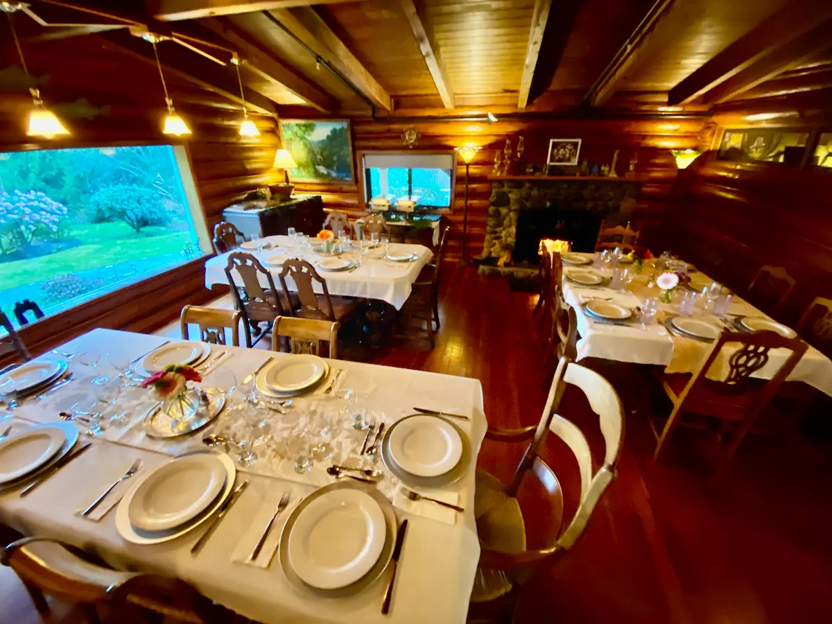 Log cabin dining hall with set banquet tables, roaring stone fireplace, and panoramic garden window at Ocean Wilderness Inn, Shirley BC